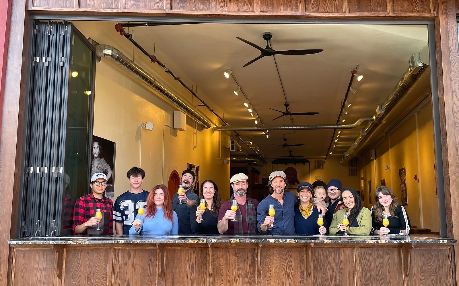 The Lumina team raising glasses of orange juice at the open bar window, the long cafe behind them