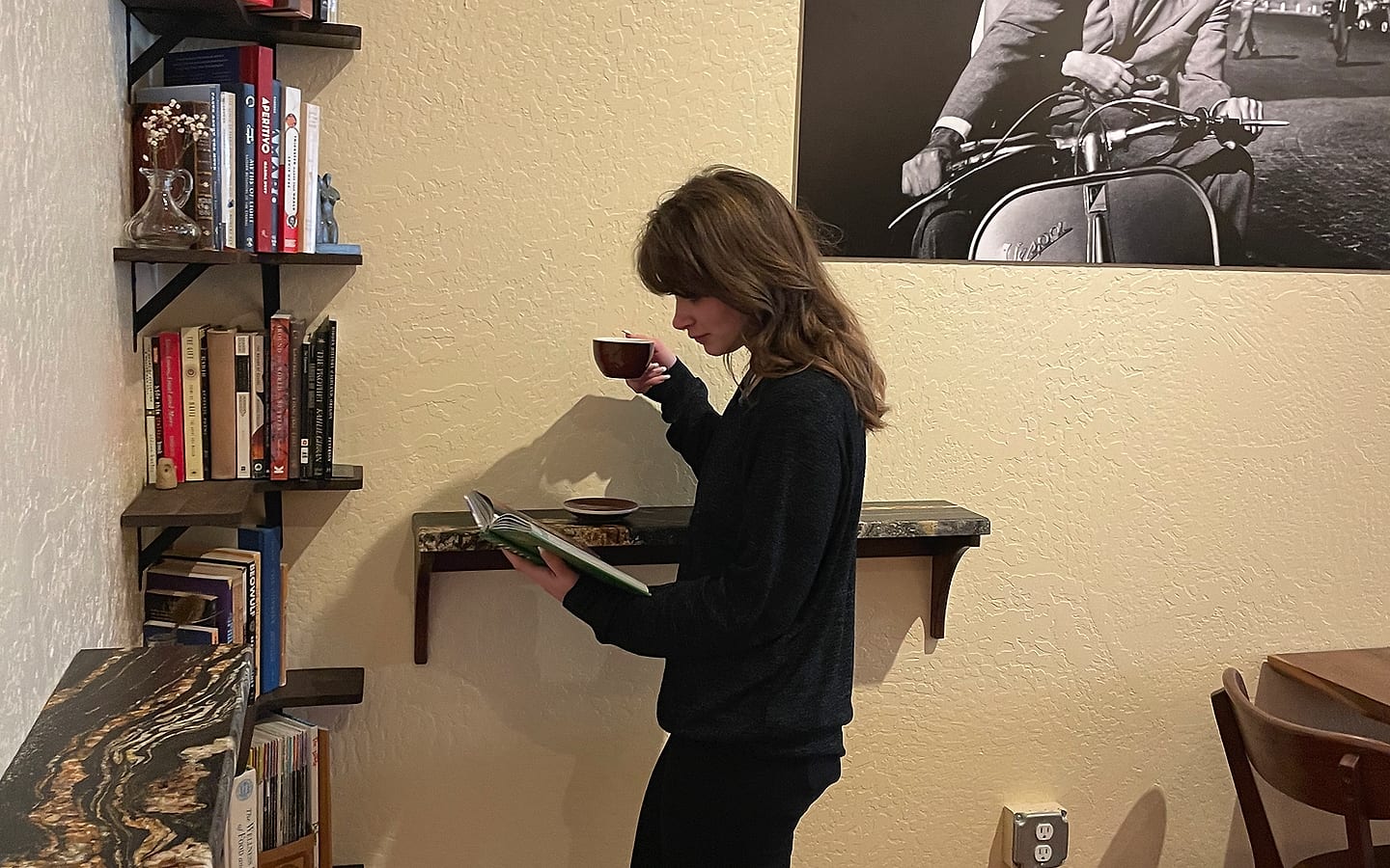 A guest sipping from an espresso cup while reading beside the cafe bookshelf