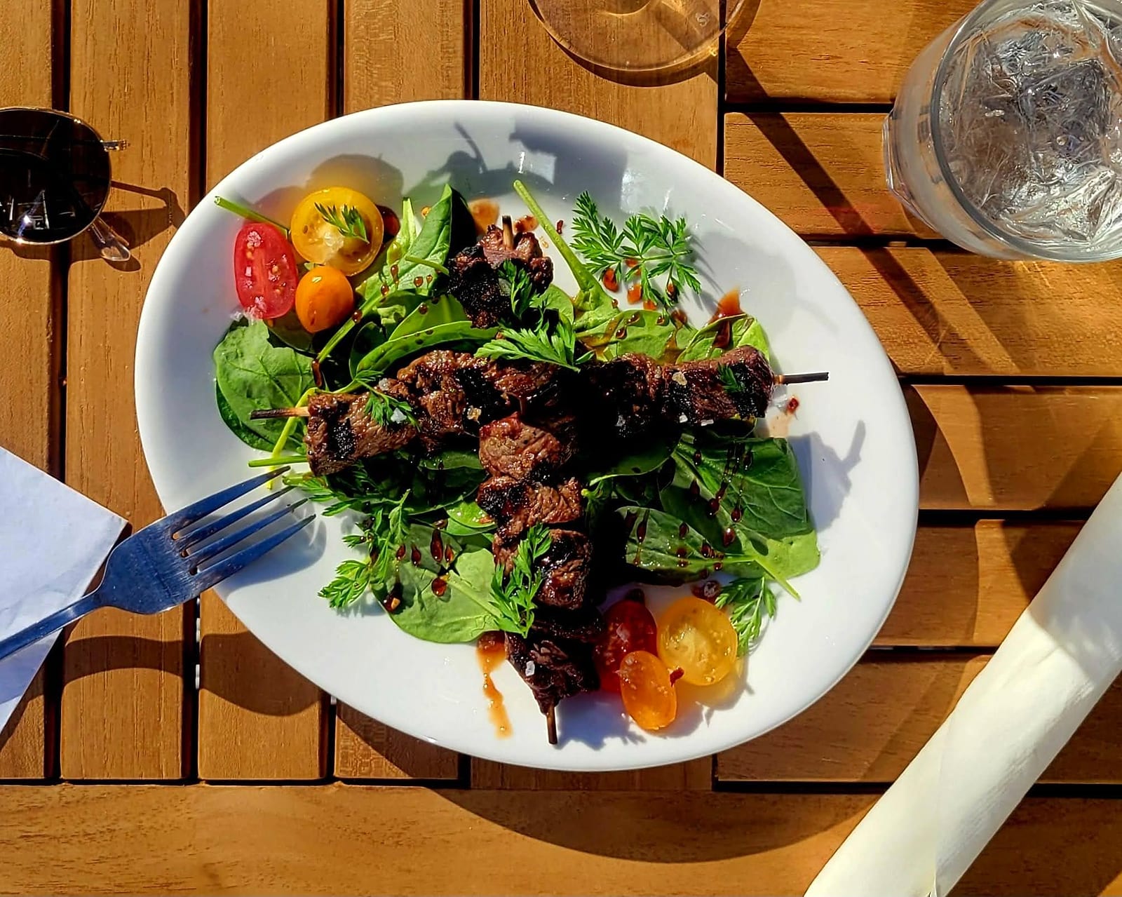 Grilled meat skewers over greens and cherry tomatoes on a wooden outdoor table
